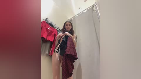 Media: Video of a young woman with long, curly hair, wearing a red plaid shirt, standing in a brightly lit dressing room. Clothes are hung on a rack to her left.