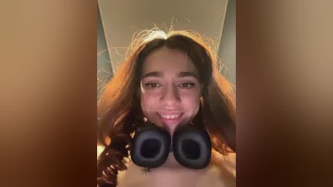 Media: Video of a young woman with medium brown skin, long wavy hair, and a bright smile. She holds large black headphones in front of her face, obscuring her eyes, in a playful manner.