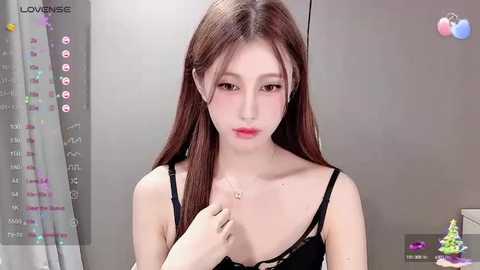 Media: Video of a young East Asian woman with long brown hair and pale skin, wearing a black lace bra, standing in a minimalist room with a grey curtain.