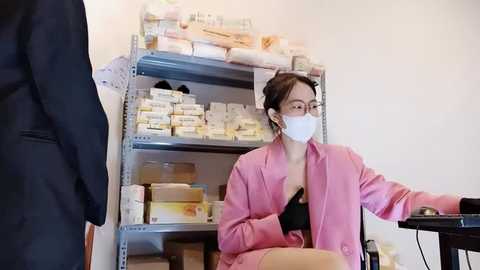 Media: Video of a woman in a pink coat, white mask, and glasses sitting at a desk, looking at a computer screen, with medical supplies on shelves in the background.