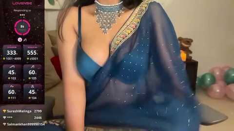 Media: Video of a South Asian woman in a blue saree with gold embroidery, wearing a statement necklace, seated indoors. The background features a pink balloon and a TV screen displaying social media statistics.