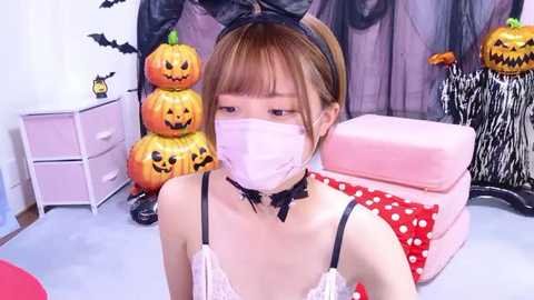 Media: Video of a young East Asian woman with fair skin and brown hair, wearing a white lace bra, black choker, and pink face mask, posing in a Halloween-themed room with jack-o'-lanterns, bats, and a polka-dotted cushion.