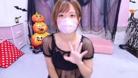 Media: Video of an Asian woman with short brown hair, wearing a black sheer dress, face mask, and black gloves, in a Halloween-themed room with carved pumpkins, bats, and a red polka-dot cushion.