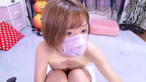 Media: A video of a nude, pale-skinned Asian woman with short brown hair, wearing a pink face mask, seated on a table in a room with red polka-dot cushions, Halloween decorations, and black lace curtains.