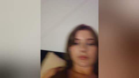 Media: Blurry video of a young woman with fair skin and long brown hair, slightly out of focus, against a beige and white background, possibly in a bedroom.