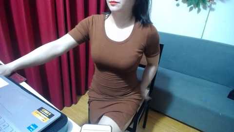 Media: A video of a fair-skinned woman with black hair, wearing a tight, brown, short-sleeved dress, sitting at a desk, adjusting a laptop. Red curtains, blue wall, wooden floor, and a chair are visible.