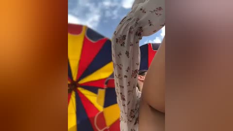 Media: Video of a person with fair skin, wearing a white floral dress, holding a large, multi-colored umbrella with red, yellow, and blue sections. The background shows a partly cloudy sky.