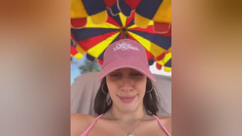 Media: Video of a woman with straight dark hair, wearing a pink baseball cap and a pink strapless top, smiling under a vibrant umbrella with red, yellow, and blue sections.