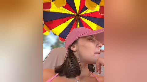 Media: A video of a woman with short brown hair, wearing a pink hat and pink top, with a colorful umbrella above her. The background is blurry, focusing on her relaxed expression.