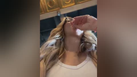 Media: Video of a blonde woman with long, wavy hair lying on her back, wearing a white shirt, with a man's hand feeding her a piece of food. Background features ornate, golden ceiling patterns.