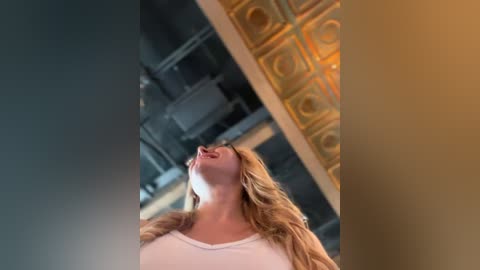 Media: Video of a young woman with long, wavy blonde hair, wearing a white tank top, smiling upward, surrounded by a modern, industrial interior with gold-patterned tiles and metallic accents.