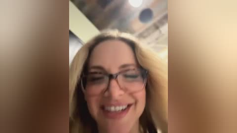 Media: Video of a blonde woman with glasses smiling, wearing a white top, in a dimly lit room with blurred, warm-colored background.