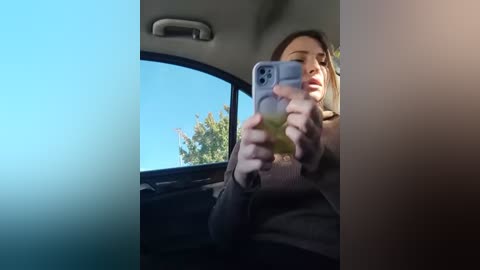 Media: A video of a woman taking a selfie through the car window, her face partially covered by her phone's camera, with a clear blue sky and green foliage visible outside.