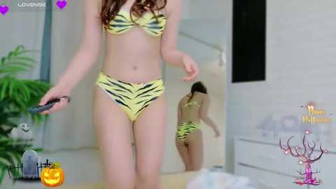 Media: Video of a fair-skinned woman in a yellow and black tiger print bikini, holding a remote, in a modern, minimalist bedroom with a mirror.