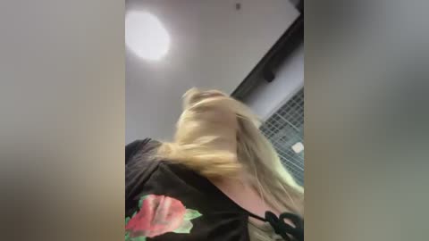 Media: A video of a blonde woman with long hair, wearing a black shirt with a red rose graphic, looking up at a bright ceiling light. The background shows a modern indoor setting with a glass partition and a white ceiling.