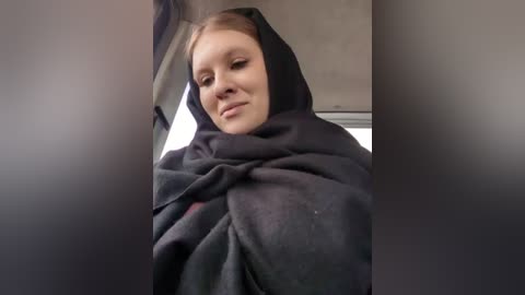 Media: A video of a young Caucasian woman with fair skin, light brown hair, and closed eyes, wearing a black hijab, seated in a car, with blurred background.