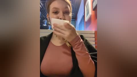 Media: A video of a Caucasian woman with light skin and blonde hair, wearing a pink long-sleeve shirt and black blazer, taking a selfie indoors, partially covering her mouth with her phone.