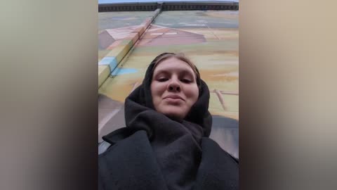 Media: Video of a young woman with fair skin, brown hair, and a serene expression, wearing a dark hooded cloak. The background features a mural with pastoral scenes, suggesting an indoor, possibly museum setting.