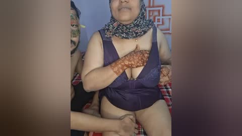 Media: Video of an Asian woman with a medium build and light skin, wearing a purple halter top exposing her large breasts, adorned with a floral-patterned headscarf. She sits on a red plaid cushion, with a man's face partially visible in the background.