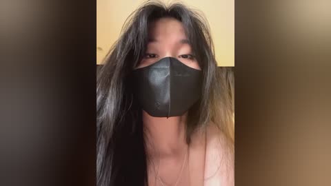 Media: Video of an Asian woman with long black hair, wearing a black face mask and light-colored top, standing against a blurred background.