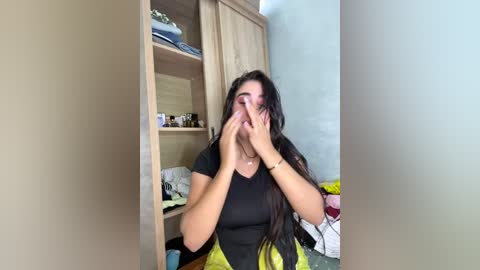 Media: A video of a young woman with long black hair, wearing a black top, covering her face with both hands in a cluttered, beige-walled bedroom with wooden shelves and a messy bed.