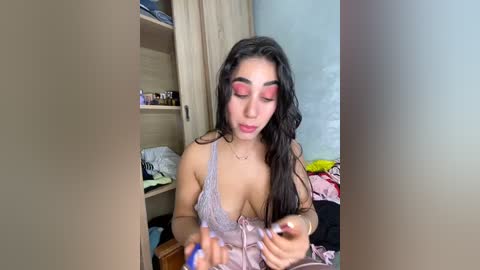 Media: Video of a young woman with long dark hair, wearing a lace bralette, applying makeup, in a messy bedroom with wooden cabinets and clothes scattered on the floor.