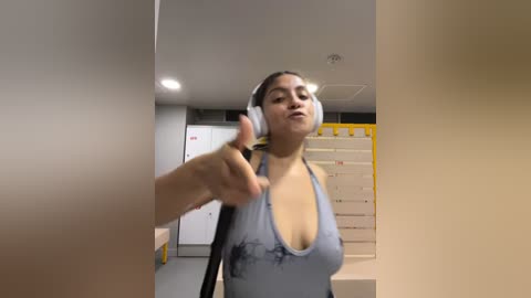 Media: A video of a woman in a modern, brightly lit room, wearing a gray halter top and white headphones. She points her index finger forward, smiling.