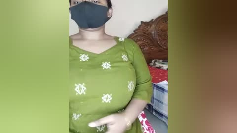 Media: Video of a South Asian woman in a green top with white floral patterns, black mask, and dark hair, standing in a bedroom with a wooden headboard, red bedspread, and blue box.