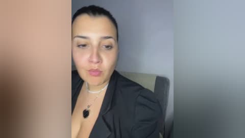 Media: A video of a light-skinned woman with dark hair and light makeup, wearing a black blazer and necklace, making a kissy face in front of a grey wall.