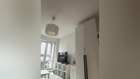 Media: A minimalist, modern apartment interior video, featuring a white room with a large window, a flat-screen TV on a white stand, and a hanging, light-colored lampshade.