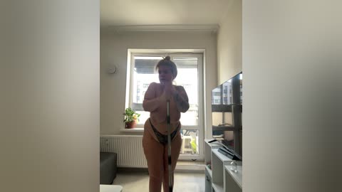 Media: Video of a nude woman with blonde hair, holding a broom, standing in a modern, well-lit apartment. She has a curvy figure and wears black panties.