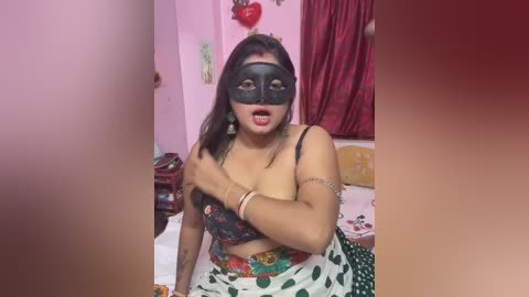 Media: Video of a South Asian woman in a black mask, floral bra, and polka-dot skirt, with a surprised expression, in a pink room with heart decorations.