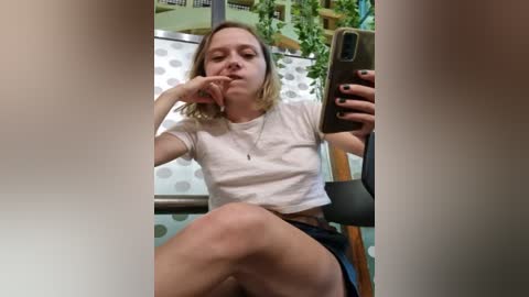 Media: Video of a young woman with shoulder-length blonde hair, wearing a white crop top and denim shorts, sitting on a chair with a green and white polka-dotted background. She holds a phone in her right hand.