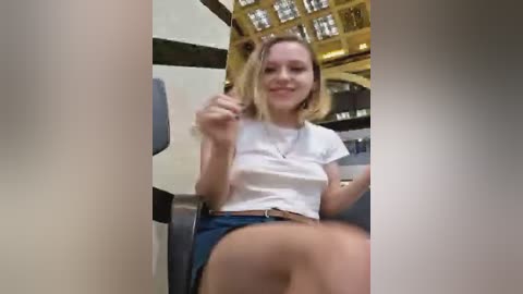 Media: A video of a young woman with shoulder-length blonde hair, wearing a white cropped top and blue shorts, seated in a modern, well-lit room with a glass ceiling.