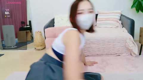 Media: Video of a young woman with a mask, wearing a white crop top and gray skirt, sitting on a bed with pink and beige linens in a cluttered room.