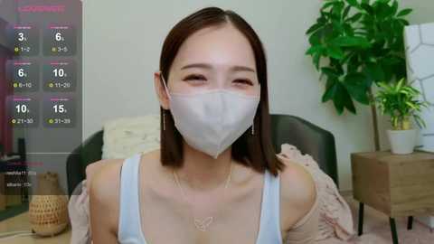 Media: A video of an East Asian woman with straight brown hair, wearing a white face mask, off-shoulder top, and a gold necklace, sitting in a modern room with a green plant and wooden nightstand.