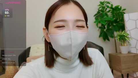 Media: Video of an East Asian woman with straight, shoulder-length brown hair, wearing a white mask, green top, and small earrings, indoors with a green plant and wooden table in the background.