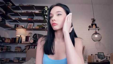 Media: Video of a pale-skinned woman with long black hair in a light blue tank top, talking on a phone, in a cozy, book-filled room with wooden shelves, a lamp, and a birdcage.