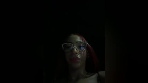 Media: A dimly lit video of a young woman with light brown skin, long red hair, and large glasses, wearing a black top, with her face partially obscured by her hand.