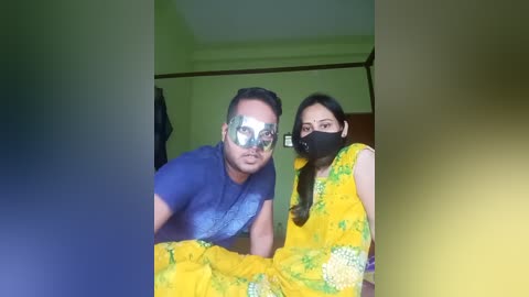 Media: Video of a man in a blue shirt and a woman in a yellow sari with floral patterns, both wearing face masks, indoors with green walls and a wooden door.