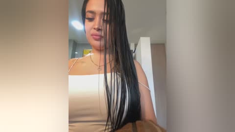 Media: A video of a young woman with long, wet black hair, wearing a beige tank top, standing indoors against a neutral background.