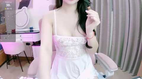 Media: Video of an Asian woman with long dark hair in a white lace dress, sitting in a modern room with a white desk, chair, and gray curtains.