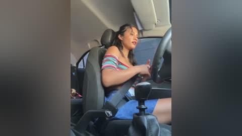 Media: Video of a woman with medium skin tone, long dark hair, wearing a striped off-shoulder top and denim shorts, sitting in a car, adjusting the steering wheel.