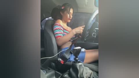 Media: Video of an Asian woman in a car, wearing a striped off-shoulder top, shorts, and a seatbelt, holding a phone, with a black bag and car seat on the floor.