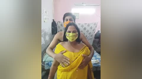 Media: A video showing a woman in a yellow saree with a mask, holding a young girl in a similar outfit. The background features a pastel-pink wall and a bed with floral-patterned bedding.