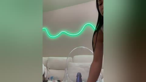 Media: A video of a woman with long black hair and a slender physique, wearing a sheer black top, standing near a white couch adorned with a neon green wave light.