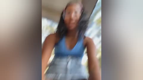 Media: A blurry video of a young Black woman with braided hair, wearing a blue tank top and high-waisted jeans, taken indoors with a window in the background.