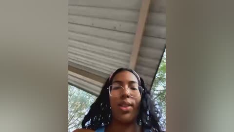 Media: Video of a young Black woman with medium brown skin and long, wavy black hair, wearing glasses, smiling, standing under a corrugated metal roof.