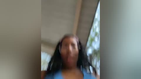 Media: A blurred, out-of-focus video of a young person with long, dark hair and a blue top, standing indoors near a window with greenery visible outside.