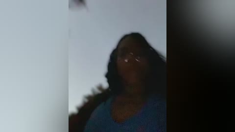 Media: A blurry video of a woman with medium-dark skin, wearing glasses and a blue sweater, standing in a dimly lit room with a white wall and a dark object in the background.